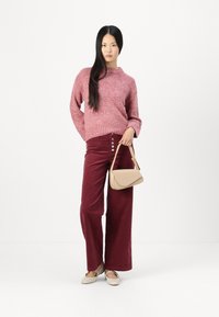 Maglione rosa a maglia con scollatura alta, abbinato a pantaloni in corduroy bordeaux a gamba larga e una borsa beige. Calzature casual completano il look.