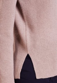 Pull en maille côtelée rose clair avec des manches longues, présentant des fentes sur les côtés au niveau de l'ourlet. La texture semble douce et tricotée avec une coupe ajustée.