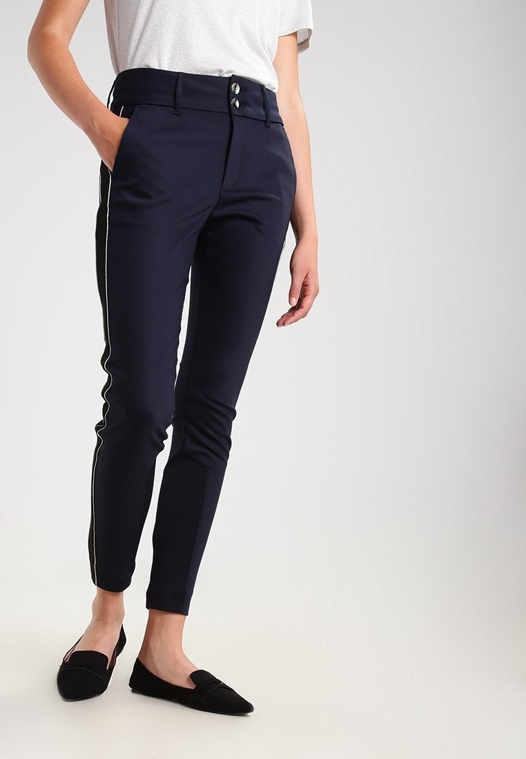 Kvinna iklädd marinblå byxor med sidostripes och en löst sittande grå topp, stående med en hand i fickan och vänd något åt vänster.