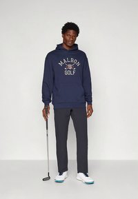 Marineblauwe hoodie met "MALBON GOLF" logo, kangoeroezak, grijze broek en witte golfschoenen met blauwe accenten; staand met een golfclub.