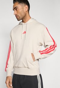 Felpa con cappuccio beige in misto cotone, caratterizzata da un design a tre strisce rosse sulle maniche e una tasca frontale a marsupio, con cappuccio regolabile con coulisse.