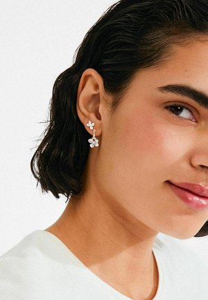 Silberne, blumenförmige Ohrringe mit Kristallakzenten, mit Hakenverschluss. Vor dem Ohr eines Models mit kurzen dunklen Haaren getragen.
