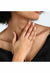 Guldring med en smal band och delikata klara ädelstenar. Bärs på en hand som vilar på nyckelbenet, mot en svart topp.