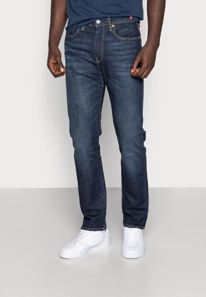 Jeans in denim blu scuro con una vestibilità dritta, leggeri segni di usura e dettagli di cucitura arancioni. Abbinati a sneaker bianche.