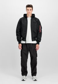 Black bomber jacket with a hood, zip front, ribbed cuffs, and a logo patch. Worn over a white t-shirt with black cargo pants and sneakers.