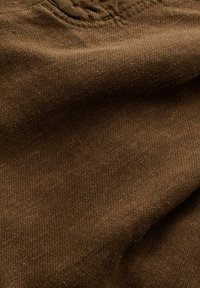 Tissu marron doux avec texture de tissage visible et plis subtils, suggérant un gros plan de matière vestimentaire.