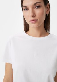 White cotton t-shirt featuring a round neckline and short sleeves, with a subtle embossed logo on the chest. Smooth texture, relaxed fit.