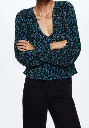 Femme les bras croisés portant un chemisier à manches longues avec un col en V profond, à motif floral bleu et vert, et un pantalon noir taille haute.