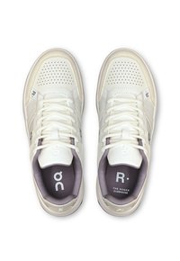 White leather sneakers featuring perforated details, white laces, and a purple inner lining. Includes branding elements on the side and tongue.