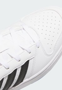 Bijela atletska cipela s crnim akcentima, koja sadrži perforirane detalje, teksturirani kožni materijal i bijele vezice.