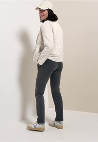 Licht beige jack met rits, grijze slim fit broek en witte sneakers met blauwe accenten. Model draagt een lichte pet, gefotografeerd van achteren.