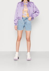 Vaatteena on purppurainen satiin bomber-takki monivärisen pyörteisen kuvion kanssa, yhdistettynä vaaleisiin denim-shortsihin ja pastellinvärisiin korkeat topit -tennikeihin.