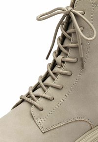 Beigenvärinen mokkanahkainen nilkkuri, jossa on tasaiset nauhat, pehmustettu kaulus ja kontrastitikkaukset sivulla; sileä pinta ja minimalistinen muotoilu.