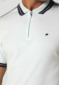 Gaiši zils kokvilnas polo krekls ar rāvējslēdzēju apkakli, tumši zilu svītru akcentiem un mazu melnu logo kreisajā krūtī.