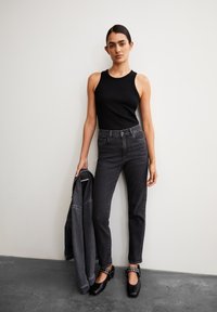 Zwarte geribbelde tanktop gecombineerd met donkergrijze high-waisted jeans. Model houdt een bijpassende denimjack vast en draagt zwarte klompen met metalen accenten.