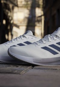 Weiße Adidas Laufschuhe mit marineblauen Streifen auf Kopfsteinpflaster, die die Cloudfoam-Dämpfung in einer urbanen Außenumgebung hervorheben.
