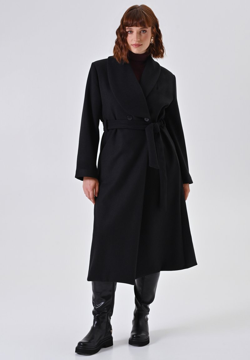 Manteau en laine noir avec une taille ceinturée, grands revers et fermeture à double bouton. Porté sur un col roulé, associé à des bottes noires jusqu'aux genoux.