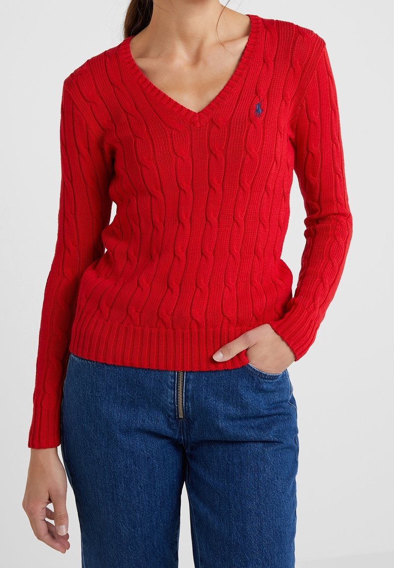 Femme portant un pull en V rouge en tricot à torsades avec des manches longues et un jean bleu avec fermeture éclaire à l'avant, main dans la poche.