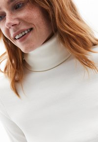 Cremfarbener Rollkragenpullover mit gerippter Textur; weicher Stoff, figurbetonte Silhouette, die einen Teil des Gesichts mit Sommersprossen und rotbraunem Haar zeigt.