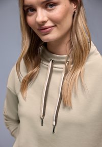 Sudadera con capucha beige de cuello alto, con cordones contrastantes en marrón y blanco. Textura lisa, diseño ceñido y sin patrones visibles.