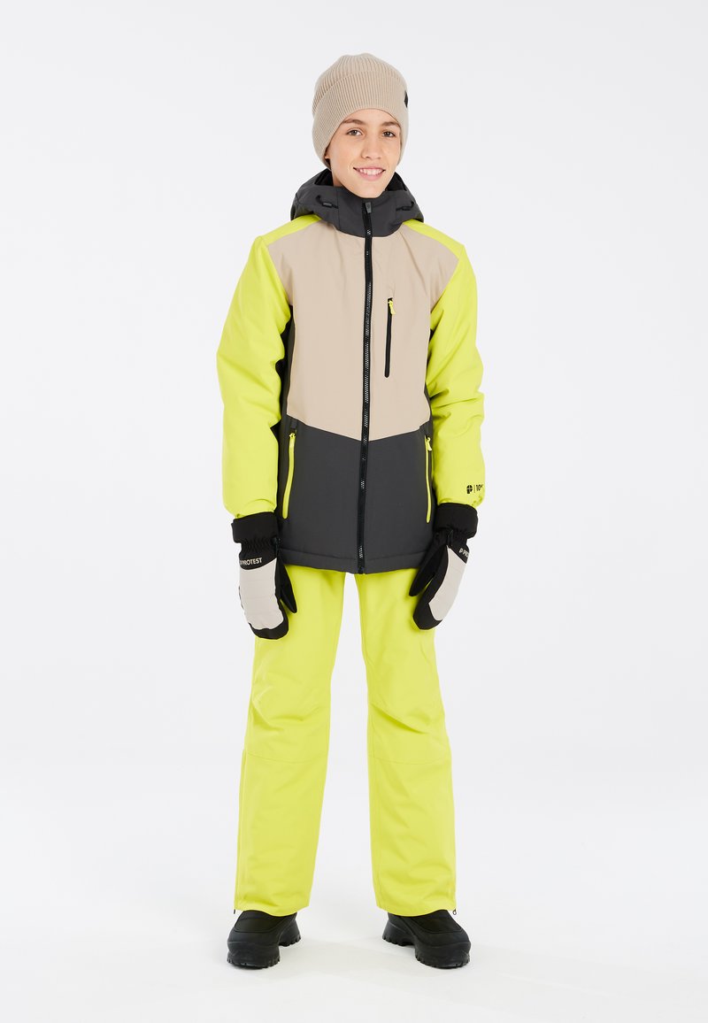 Heldere gele sneeuwjas met grijze en beige accenten; bijpassende gele broek; zwarte handschoenen; gebreide beige beanie; stevige zwarte laarzen.