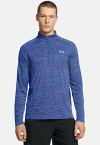 Blaues langärmeliges Sportshirt mit einem Viertelreißverschluss, das aus einem strukturierten Stoff besteht und ein hellblaues Logo auf der Brust trägt.