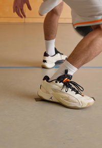Person iført hvide og sorte sneakers samt hvide sokker, der dribbler en basketball indendørs på et gymgulv.