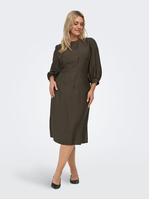 Femme aux cheveux blonds portant une robe vert olive longueur genou avec des manches bouffantes trois-quarts, debout, jambes croisées, avec des talons noirs.
