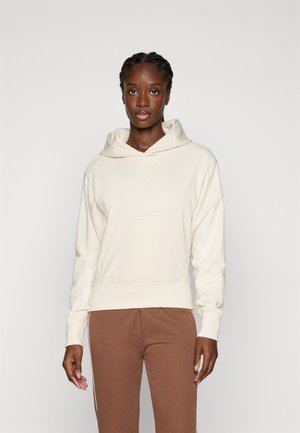 Joven mujer con el pelo trenzado, vistiendo una sudadera con capucha de color crema y pantalones marrones, de pie contra un fondo claro y liso.