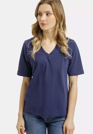 Kobieta o falowanych blond włosach, ubrana w granatowy T-shirt z dekoltem w serek i niebieskie dżinsy, spoglądająca w lewo na jednolitym tle.