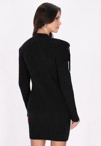 Schwarzes geripptes Strickkleid mit langen Ärmeln und figurbetontem Schnitt. Mit Fransenakzenten an den Schultern. Weiche Textur, knielange Länge.