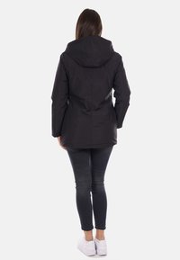 Giacca nera impermeabile con cappuccio, caratterizzata da una texture liscia e un design leggermente aderente, abbinata a jeans scuri e sneakers bianche.