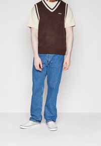 Brown knitted vest with white trim, paired with a light beige t-shirt, blue straight-leg jeans, and white checkered slip-on shoes.