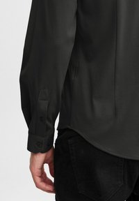 Dark gray long-sleeve shirt with a smooth texture, featuring buttoned cuffs and angled cuff details. Worn with black denim pants.
