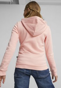 Sudadera de color rosa claro con capucha, puños y bajo acanalados, combinada con jeans de color azul oscuro. Textura suave, corte casual y detalles de costura visibles.