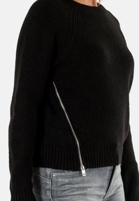 Suéter negro acanalado de material grueso, con detalle de cremallera plateada en el lado, con un cuello alto y un diseño ajustado.
