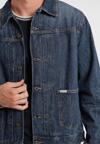 Giubbotto di jeans blu scuro con chiusura a bottoni, dettagli in metallo e due tasche laterali. Presenta cuciture a vista e un'etichetta con il marchio.