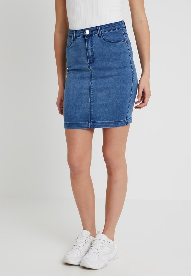 Femme portant une mini-jupe en denim bleu avec bouton et fermeture éclair à l'avant, associée à des baskets blanches et un top blanc sans manches.