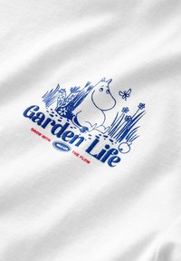 Vit bomullströja med blått grafiskt tryck av en karaktär bland blommor och gräs, med texten "Garden Life" och "Grow with the flow."