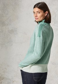 Sweter dziany w miętowym zielonym kolorze z prążkowaną fakturą, wysokim kołnierzem i kontrastującym jaśniejszym wykończeniem. Połączony z ciemnymi dżinsami.