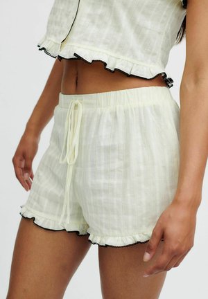 Femme portant un short texturé couleur crème avec cordon de serrage et bords froncés bordés de noir, associé à un top court assorti.