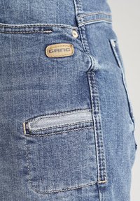 Farkut, joissa on sininen pesty pinta, takatasku jossa vaakasuora tikkaus, ja nahkaleima, jossa lukee "GANG."