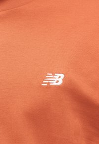 Πορτοκαλί βαμβακερό T-shirt με λευκό κεντημένο λογότυπο "NB" στην αριστερή πλευρά του στήθους. Λείο ύφασμα και στρογγυλή λαιμόκοψη.