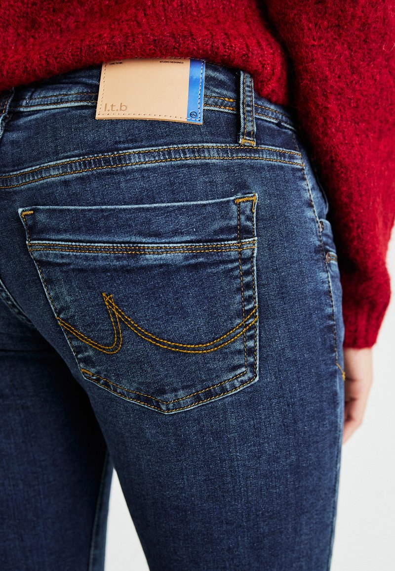 Blaue Jeans aus hochwertigem Denim mit gelber Naht, fünf Taschen und einem braunen Lederlabel an der Taille. Der Stoff hat eine glatte Textur.