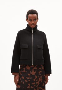Chaqueta corta negra hecha de tela texturizada, con cuello alto, cremallera frontal y dos bolsillos en el pecho. Combinada con una falda estampada.