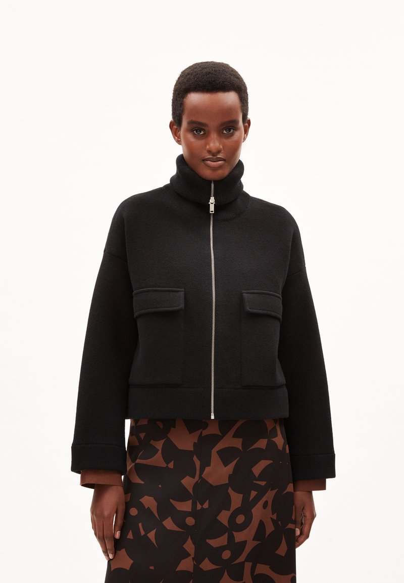Chaqueta corta negra hecha de tela texturizada, con cuello alto, cremallera frontal y dos bolsillos en el pecho. Combinada con una falda estampada.