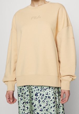 Beige FILA-sweatshirt met lange mouwen, gedragen over een groen-blauw bloemenpatroongebloemde rok, getoond van de nek tot halverwege de dij.