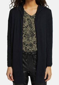 Black, hooded cardigan made of lightweight material, featuring a front opening and side pockets, worn over a green, patterned top.