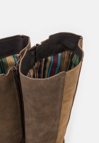 Botas marrones de ante con una ligera textura, con cierre de cremallera y un forro interior de tela a rayas coloridas. El borde superior es festoneado.