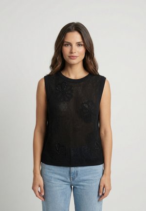 Donna con lunghi capelli castani che indossa una canotta nera in rete senza maniche con motivi floreali e jeans azzurri, in piedi davanti a uno sfondo grigio uniforme.